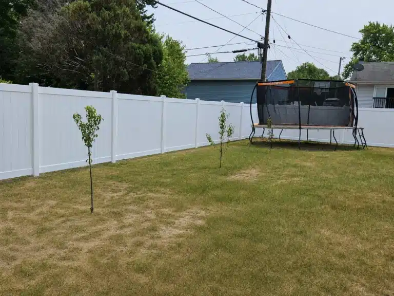 White vinyl privacy fence surrounding a backyard with a trampoline and young trees in Pasadena, MD.